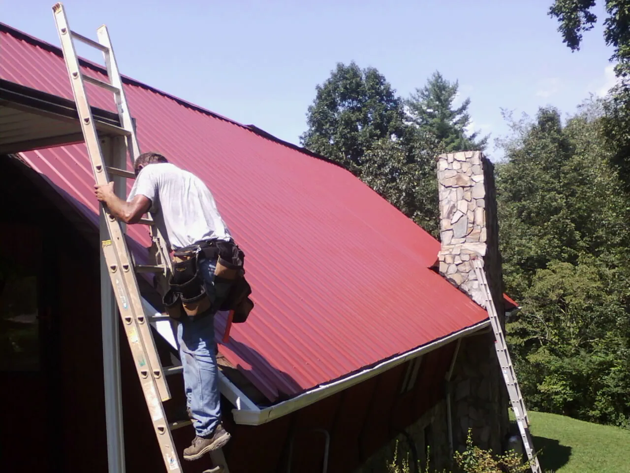 Roofer on a ladder beside a red metal roof and stone chimney — Soffit & Fascia Repair by Roofing Brothers in Grandview